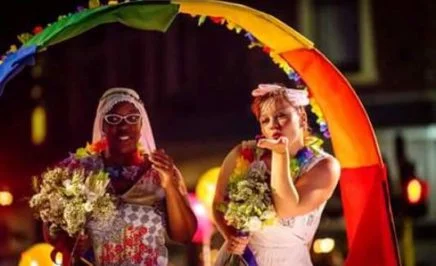 Image from Pride Rally Perth 2017. Two brides holding hands, standing under a rainbow arch. One bride is blowing a kiss to the crowd while the other holds a bouquet.