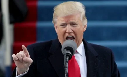 A photograph of President Donald Trump at his inauguration in January 2017. The image shows President Trump standing in front of a microphone in mid-sentence and pointing the index finger on his right hand towards the crowd (out of picture). Trump is wearing a dark coat, white shirt and red tie.