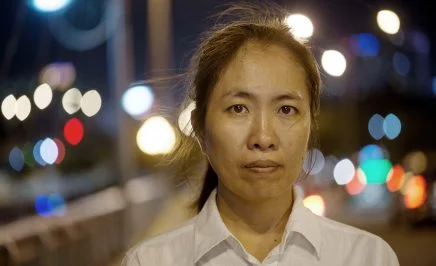 A Vietnamese women wearing a white shirt stares into the camera. It is nighttime and the lights of a city twinkle behind her.