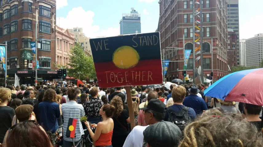 People gather to protest for Indigenous rights. Someone holds a sign that says: 'We stand together'.