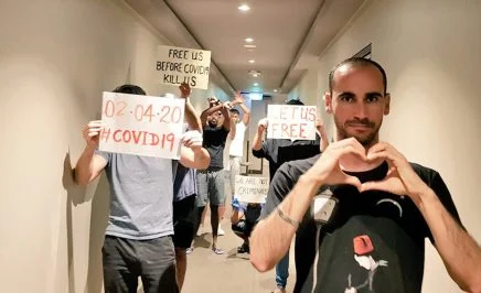 A small group of men stand in a hotel hallway, holding handmade signs asking 'Free us before COVID19 kills us'. One man creates heart symbol with his hands.