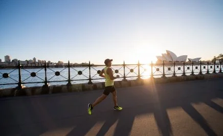 A man jogs along the harbour in Sydney, the sun low on the horizon and the Sydney Opera House in the background.