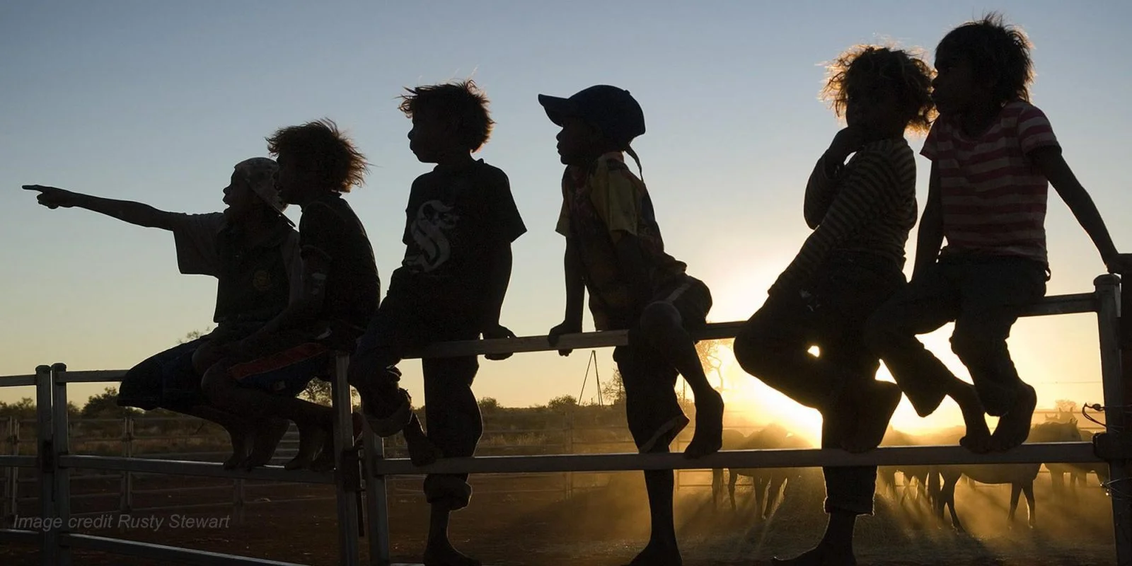 Indigenous children are silhouetted against the sunset as they sit on a fence