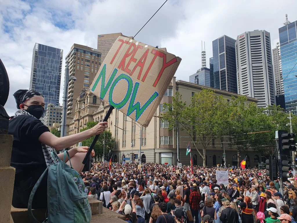 A protestor holds a sign with the words TREATY NOW in front of a crowd of demonstrators at Parliament House in Naarm (Melbourne).